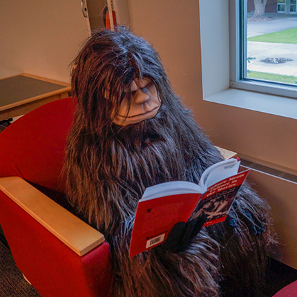 Skitch the Sasquatch reading a book while seated in a red chair