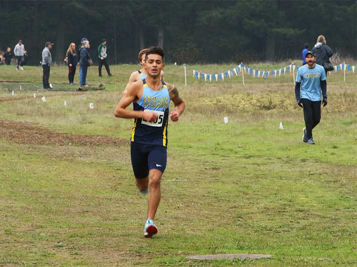 Men's cross country team running on grass with coach in the background